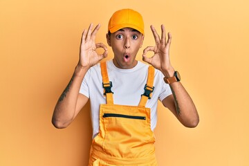 Young handsome african american man wearing handyman uniform over yellow background looking surprised and shocked doing ok approval symbol with fingers. crazy expression