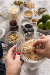 Woman hands taking pieces of oat grains in a glass jar. In the background there are some ingredients for a granola recipe.