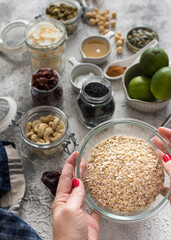Glass jar full of oat cereal held by woman hands surrounded by granola ingredients in bowls.