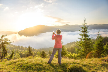 The girl photographs the sunrise in the mountains. A tourist girl at the top of the mountain photographs the rising sky.