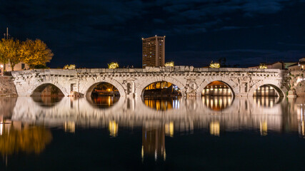 Ponte romano antico sopra al fiume di notte con grattacielo sullo sfondo e luci situato a Rimini - obrazy, fototapety, plakaty
