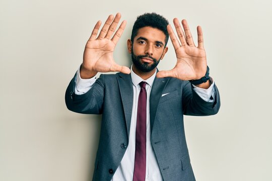 Handsome hispanic man with beard wearing business suit and tie doing frame using hands palms and fingers, camera perspective