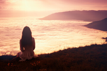 Romantic beautiful young woman in a white dress sitting on a mountain top above the clouds at...