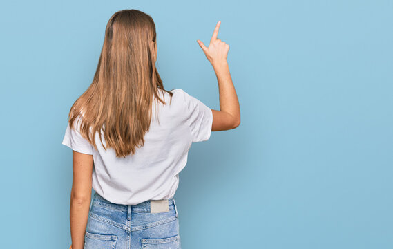 Beautiful young blonde woman wearing casual white t shirt posing backwards pointing ahead with finger hand