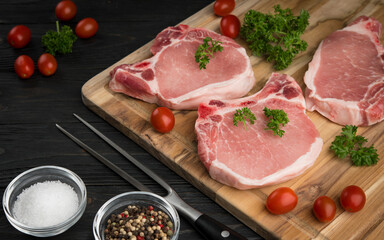 Meat steaks. Raw pork or beef ribeye meat with bone. Ingredients for barbecue. Cherry tomatoes, colored pepper, sea salt and parsley on black wooden background. Veal chunk. Food photography.