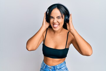 Naklejka premium Young beautiful hispanic girl listening to music using headphones sticking tongue out happy with funny expression.