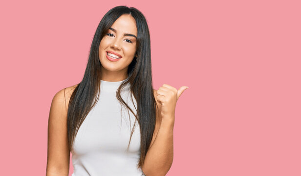 Young beautiful hispanic girl wearing casual clothes smiling with happy face looking and pointing to the side with thumb up.