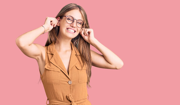 Young beautiful blonde woman wearing casual clothes and glasses smiling pulling ears with fingers, funny gesture. audition problem