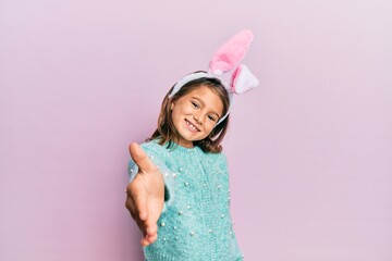 Little beautiful girl wearing cute easter bunny ears smiling friendly offering handshake as...