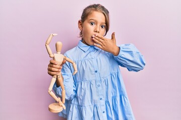 Little beautiful girl holding small wooden manikin covering mouth with hand, shocked and afraid for mistake. surprised expression