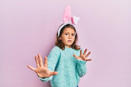 Little beautiful girl wearing cute easter bunny ears afraid and terrified with fear expression stop gesture with hands, shouting in shock. panic concept.