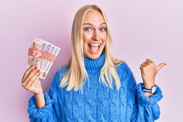 Young blonde girl holding hong kong 10 dollars banknotes pointing thumb up to the side smiling happy with open mouth