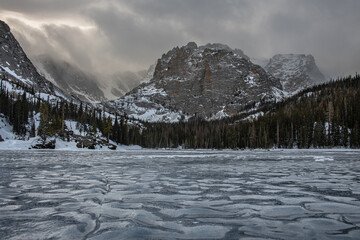 View of Loch Lake on the Glacier Gorge Trail in Rocky Mountain National Park Colorado in winter. A mountain can be seen with a frozen over lake, water ripples, snow, and dark clouds. 