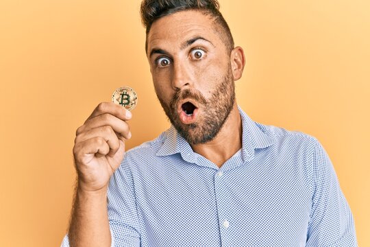 Handsome man with beard holding virtual currency bitcoin scared and amazed with open mouth for surprise, disbelief face