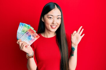 Young chinese woman holding swiss franc banknotes doing ok sign with fingers, smiling friendly gesturing excellent symbol