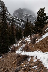 View of a mountain peak between a valley in the winter in Rocky Mountain National Park in Colorado. Other peaks can be seen along with a forrest of trees and a cloudy sky.