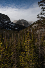 View of a mountain peak between a valley in the winter in Rocky Mountain National Park in Colorado. Other peaks can be seen along with a forrest of trees and a cloudy sky.
