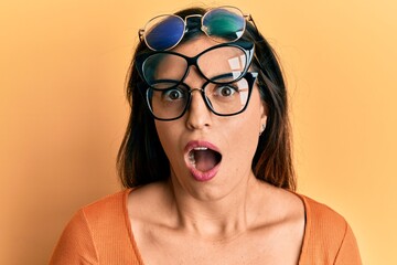 Beautiful hispanic woman wearing many glass over face scared and amazed with open mouth for...