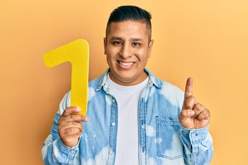 Young latin man holding number one smiling with an idea or question pointing finger with happy...