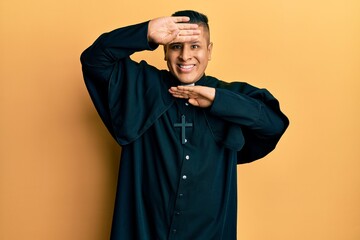 Young latin priest man standing over yellow background smiling cheerful playing peek a boo with...