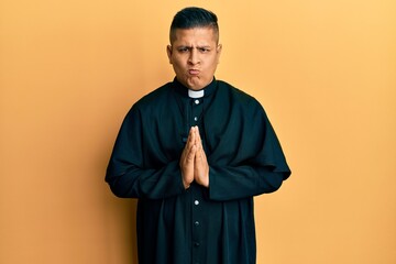 Young latin priest man praying with hands together skeptic and nervous, frowning upset because of...