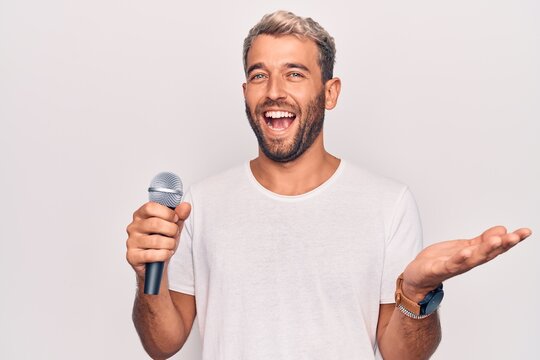 Handsome blond singer man with beard singing song using microphone over white background celebrating achievement with happy smile and winner expression with raised hand