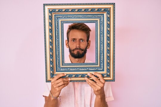 Handsome caucasian man with beard holding empty frame skeptic and nervous, frowning upset because of problem. negative person.