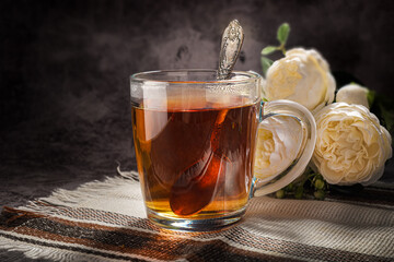 Tea is fragrant black, in a transparent mug, on a light brown napkin, with a snail bun with cinnamon and flowers in the background on a gray background