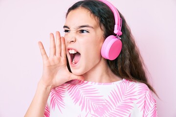 Fototapeta premium Cute hispanic child girl listening to music using headphones shouting and screaming loud to side with hand on mouth. communication concept.