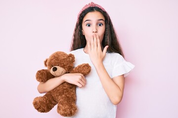 Cute hispanic child girl holding teddy bear covering mouth with hand, shocked and afraid for...