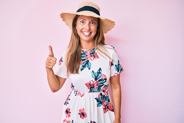 Middle age hispanic woman wearing summer hat smiling happy and positive, thumb up doing excellent and approval sign