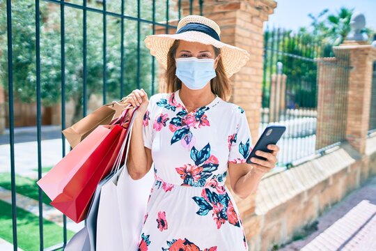 Middle age woman wearing medical mask holding shopping bags and smartphone at the city