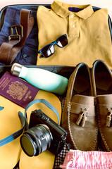 Photograph in portrait format of a suitcase packed with shorts, polo shirt, flip flops, gentlemans suede loafers, camera, sunglasses and sun cream