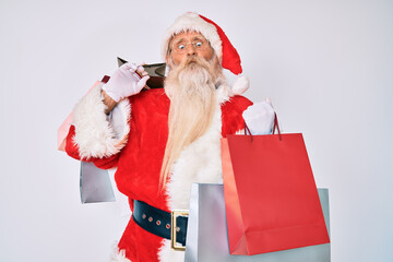 Old senior man with grey hair and long beard wearing santa claus costume holding shopping bags making fish face with mouth and squinting eyes, crazy and comical.