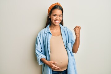 Beautiful hispanic woman expecting a baby, touching pregnant belly very happy and excited doing winner gesture with arms raised, smiling and screaming for success. celebration concept.