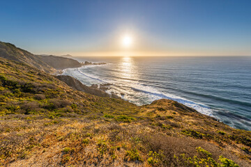 An awe sunset in an idyllic beach landscape with the sunset over the water and the sunlight generating a moody atmosphere. An amazing wil scenery with the cliffs and the sea waves splashing water