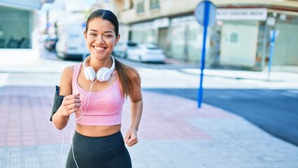 Young beautiful hispanic sport woman wearing runner outfit and headphones, smiling happy running at the town