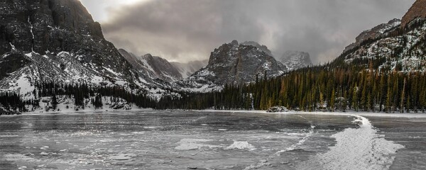 Panorama at Loch Lake on the Glacier Gorge Trail in Rocky Mountain National Park Colorado in winter. A mountain can be seen with a frozen over lake, snow, and dark clouds. 