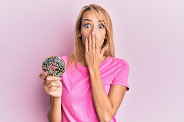 Beautiful blonde woman holding donut covering mouth with hand, shocked and afraid for mistake....