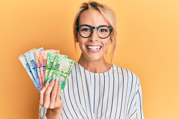 Beautiful blonde woman holding south african rand banknotes looking positive and happy standing and smiling with a confident smile showing teeth