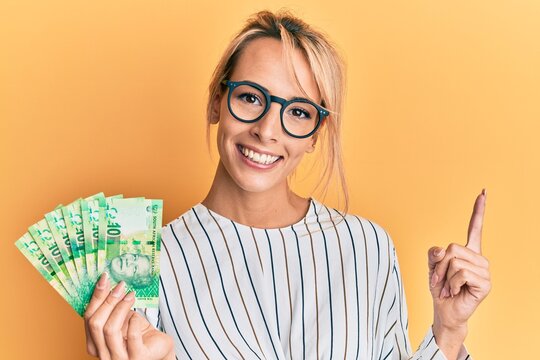 Beautiful blonde woman holding south african 10 rand banknotes smiling happy pointing with hand and finger to the side
