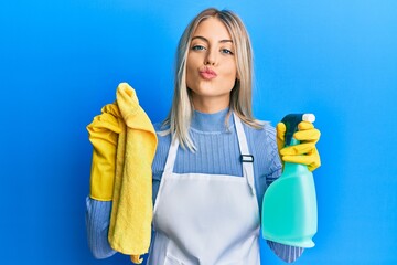 Beautiful blonde woman wearing cleaner apron holding cleaning products looking at the camera...