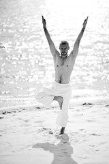 Obraz premium Young caucasian male practicing yoga in white pants on a sandy shore.
