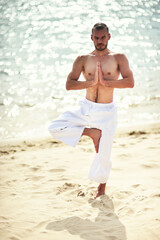 Young caucasian male practicing yoga in white pants on a sandy shore.