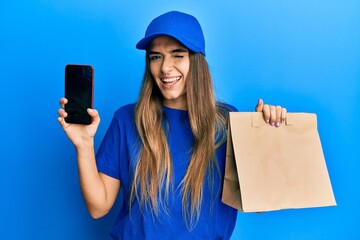 Young hispanic woman holding delivery paper bag and showing smartphone screen winking looking at...