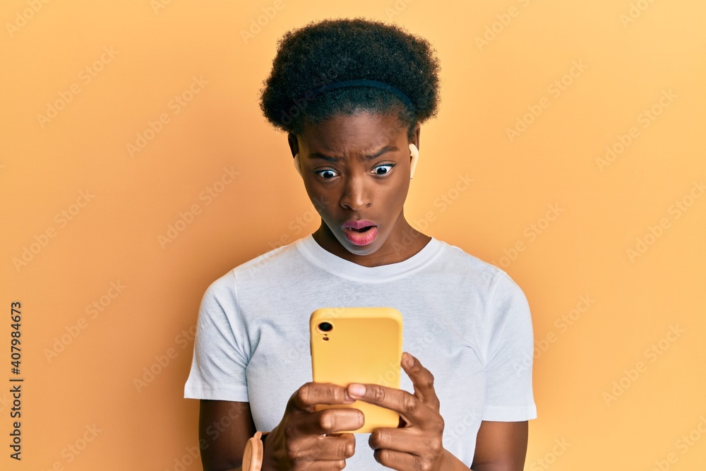 Canvas Prints Young african american girl using smartphone and earphones in shock face, looking skeptical and sarcastic, surprised with open mouth