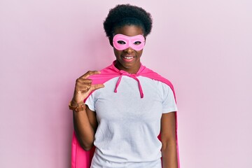 Young african american girl wearing superhero mask and cape costume smiling and confident gesturing with hand doing small size sign with fingers looking and the camera. measure concept.