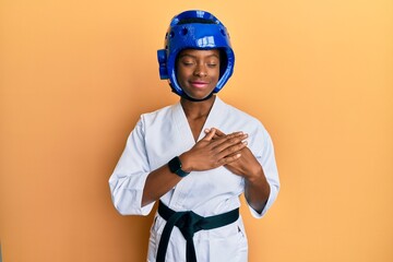 Young african american girl wearing taekwondo kimono and protection helmet smiling with hands on chest, eyes closed with grateful gesture on face. health concept.