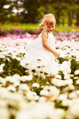 Obraz na płótnie Canvas Little blond child girl in a white dress walking between flowers in a flower bed in the park.