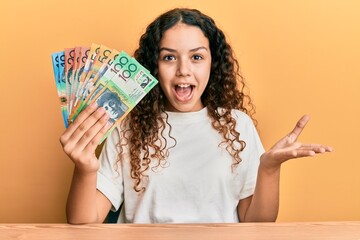 Teenager hispanic girl holding australian dollars celebrating achievement with happy smile and winner expression with raised hand